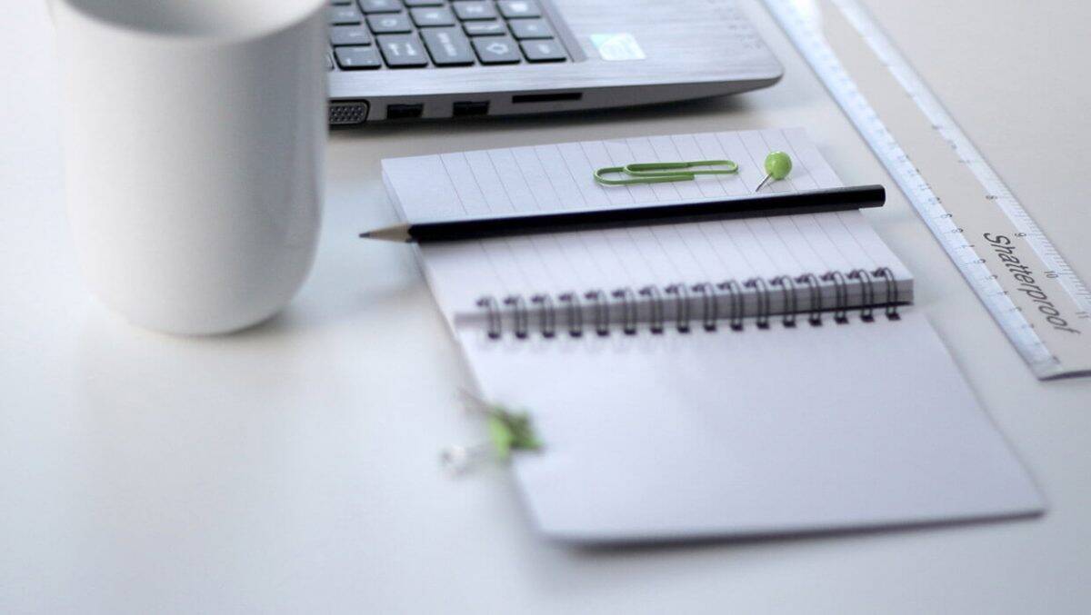 Mesa branca com notebook, caneca, caderno de espiral aberto, lápis preto, régua e clipes de papel verdes. Foco no caderno.