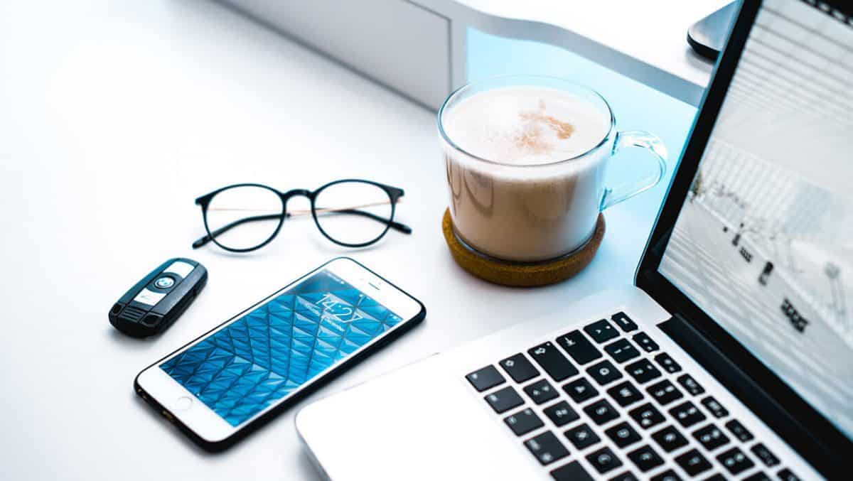 Notebook aberto ao lado de smartphone, óculos, chave de carro e uma xícara de café sobre mesa branca em ambiente iluminado.
