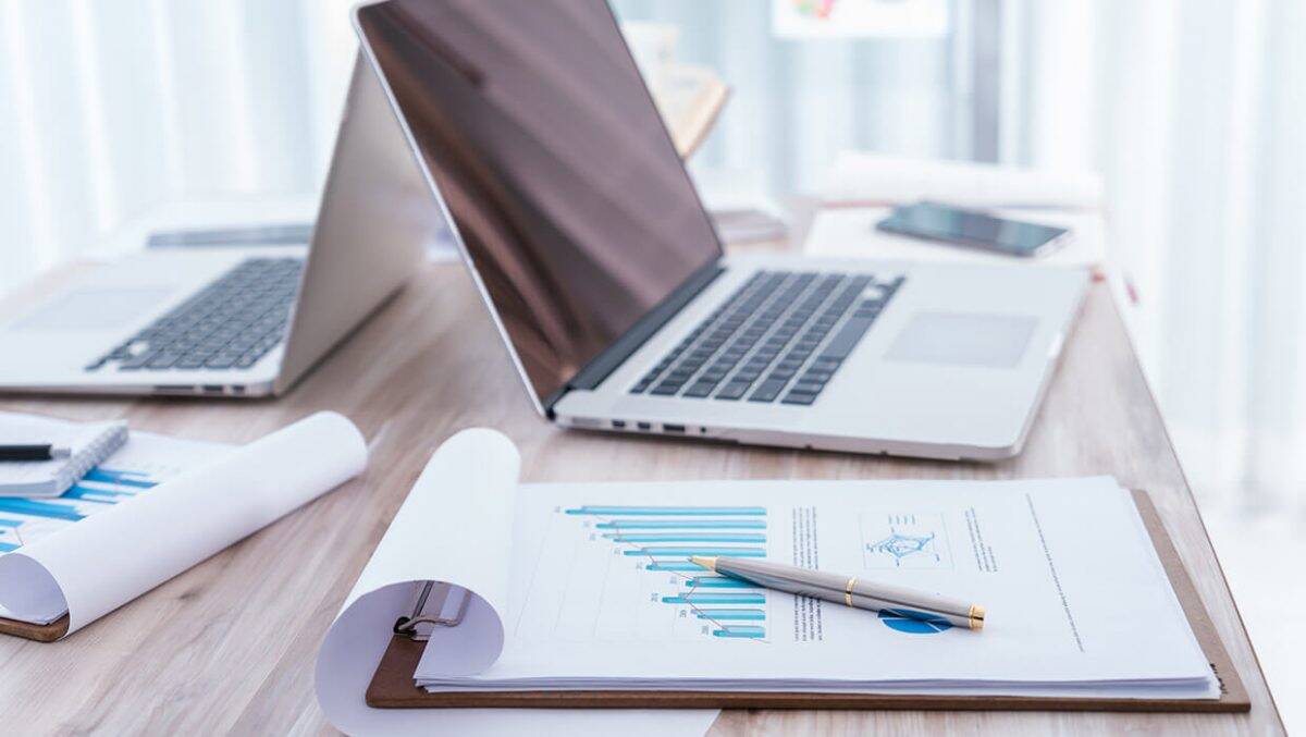 Prancheta com gráficos financeiros em primeiro plano, caneta sobre o papel e notebooks abertos em uma mesa de escritório.