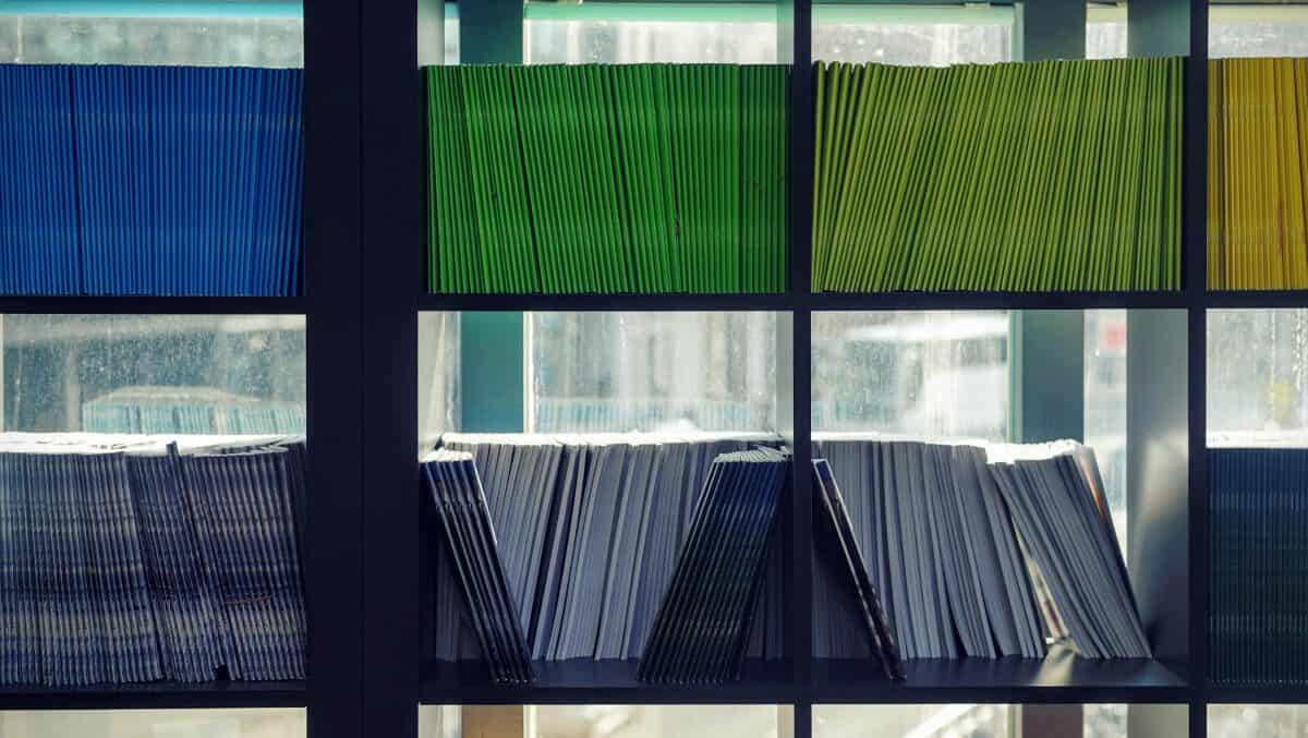 Estante com nichos quadrados contendo pastas organizadas por cores: azul, verde e amarela acima, e brancas na parte inferior.